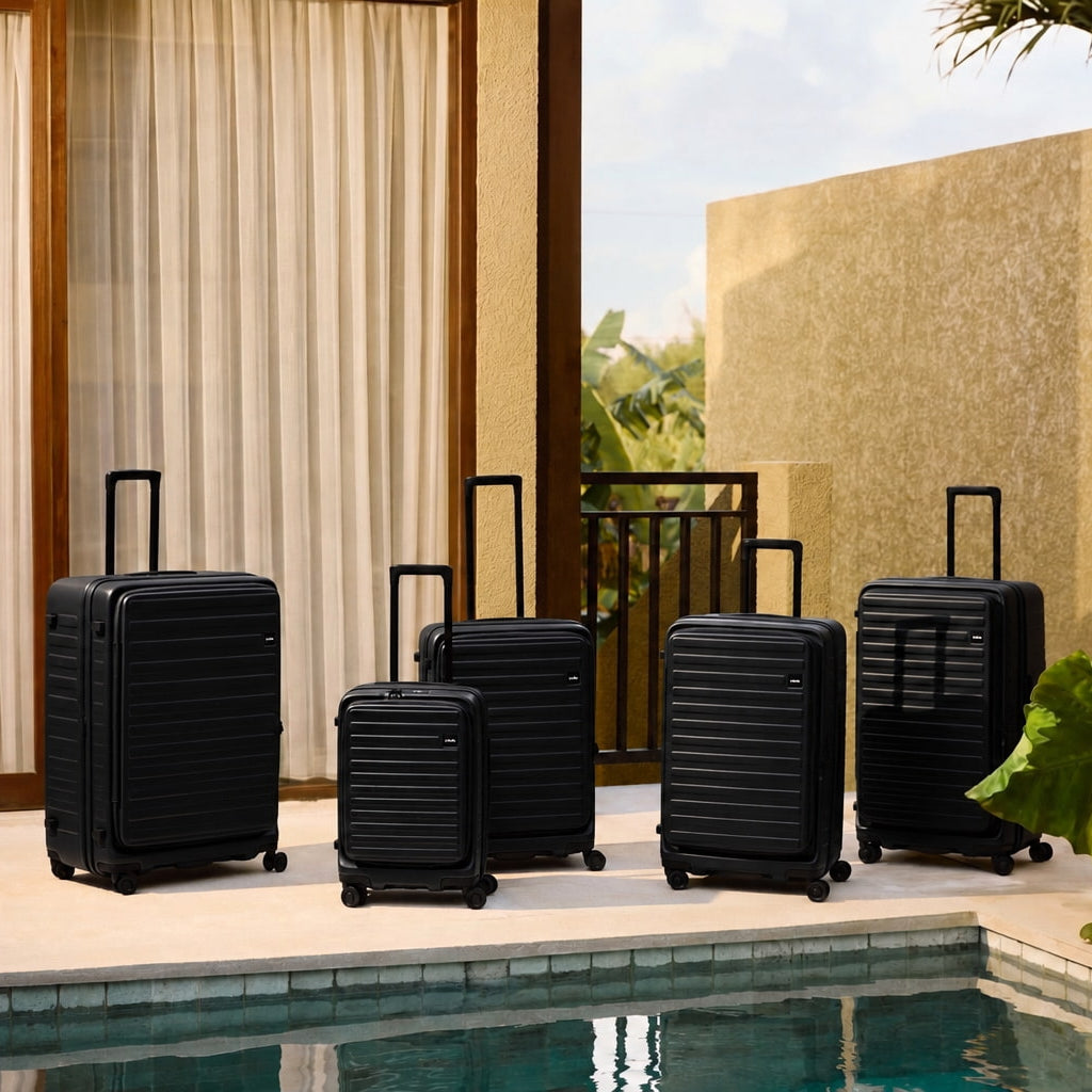 Five black hard-shell spinner suitcases in different sizes arranged on a patio beside a swimming pool, with curtains, a stucco wall, and large green leaves in the background.
